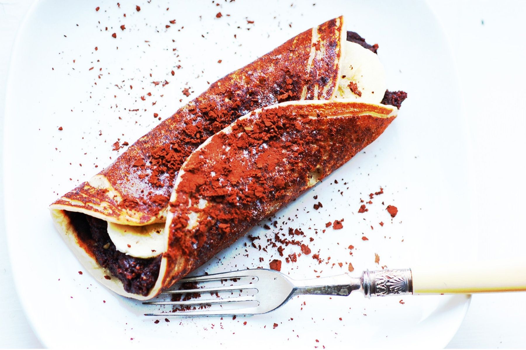 Banana pancake stuffed with bananas and vegan hazelnut spread and dusted with cocoa on a white plate